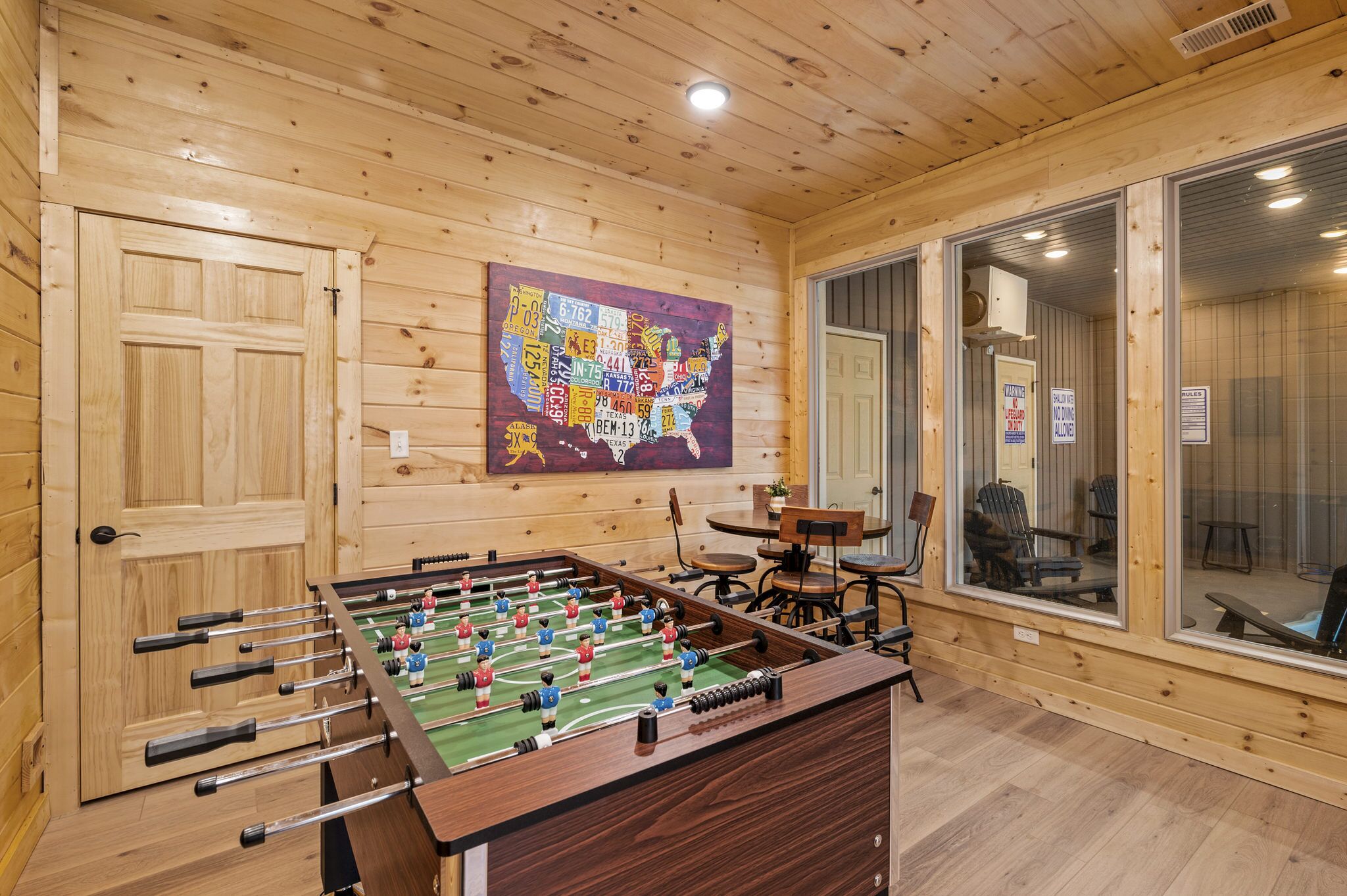A wooden-floored room with a foosball table, a map on the wall, and a sliding glass door leading to a balcony.
