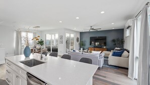 A modern kitchen with a central island, a dining area with a table and chairs, a living room with a sofa and TV, and a ceiling fan.