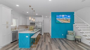 A modern kitchen with white cabinetry, a central island with bar stools, a stainless steel refrigerator, and a teal accent wall with a cityscape painting. - New Beautiful Home @ Ocean Lakes Family  Campground! BE THE FIRST TO RENT! (Myrtle Beach)