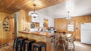 A kitchen with wooden walls, a central island with stools, a dining table with chairs, and a refrigerator.