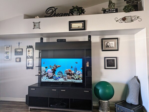 A living room with a flat-screen TV on a black entertainment unit, surrounded by various framed pictures and motivational signs on the walls.