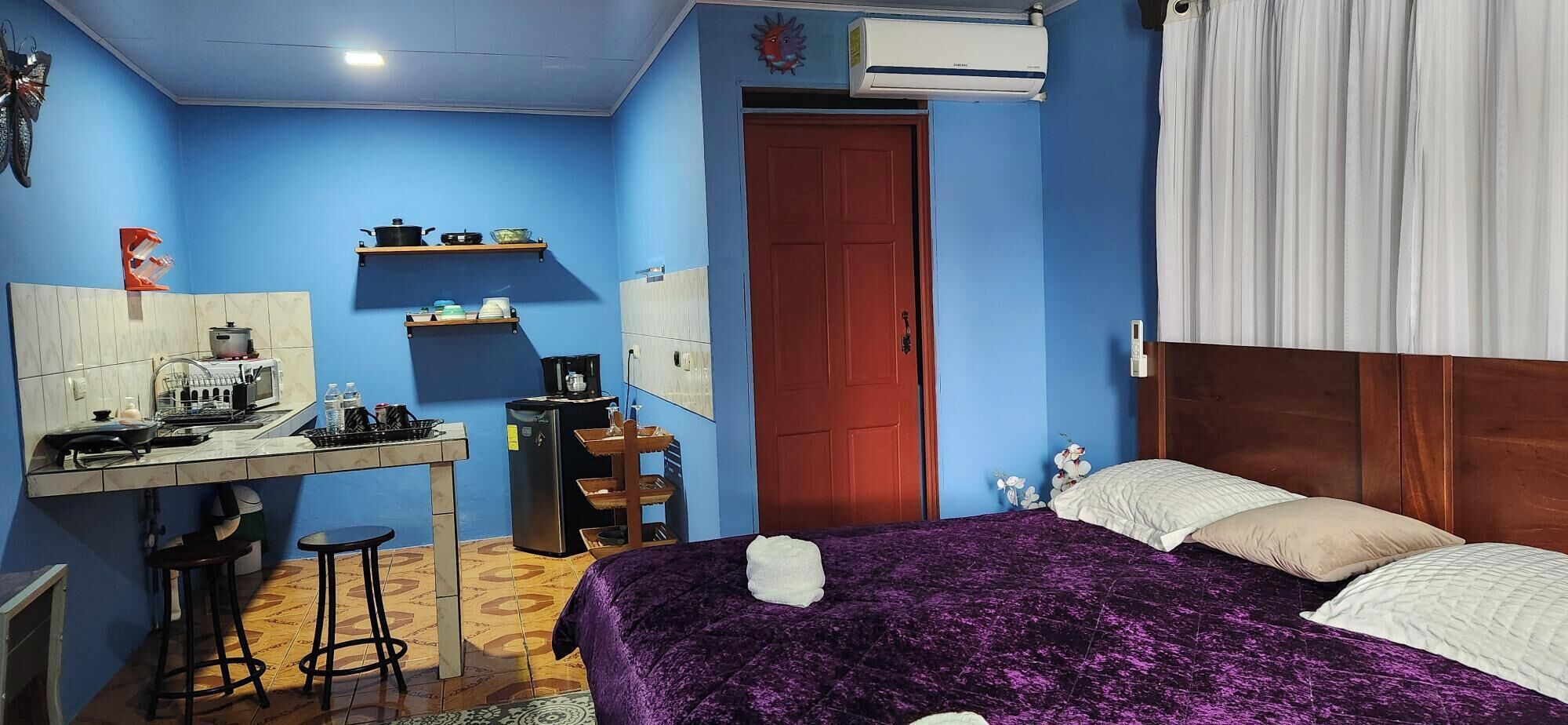 Ein Schlafzimmer mit Bett, Schreibtisch mit Stuhl, Kühlschrank, Waschbecken und einer roten Tür.