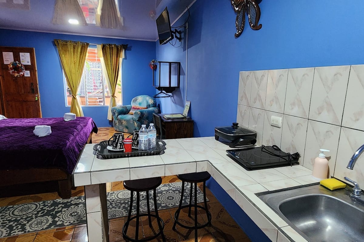 A compact hotel room with a bed, a small kitchenette, a sink, and a chair.