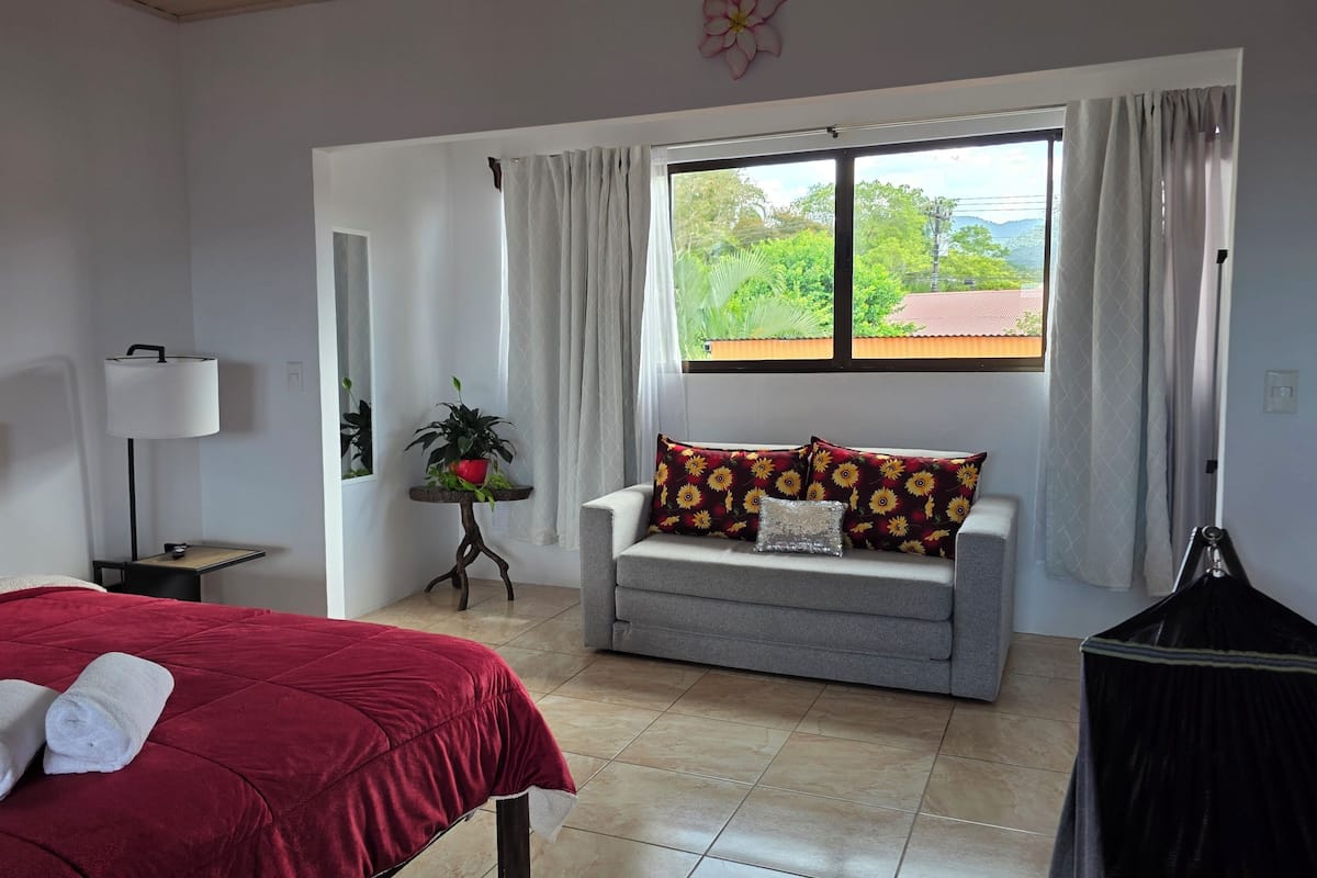 A bedroom with a bed, a sofa, a window with curtains, a lamp, and a potted plant.