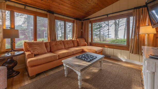 A cozy cabin interior with a sectional sofa, a wooden coffee table, and large windows offering a view of trees.