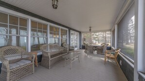 A covered porch with wicker furniture, large windows, and a view of trees.