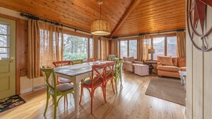 A cozy cabin interior with a wooden dining table, colorful chairs, a sofa, and a lamp.