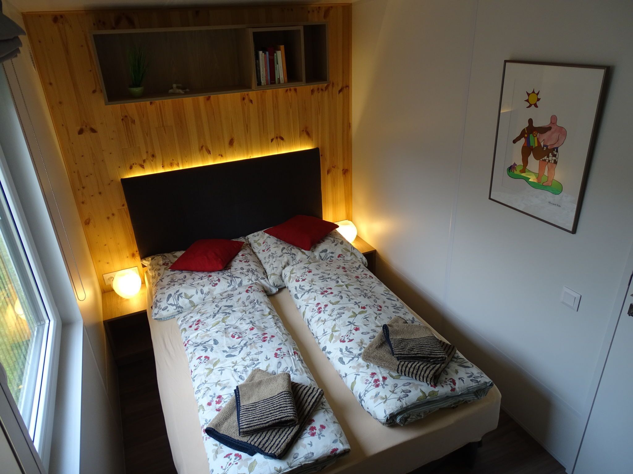 A single bed with floral bedding, two red pillows, and a headboard with built-in shelves.