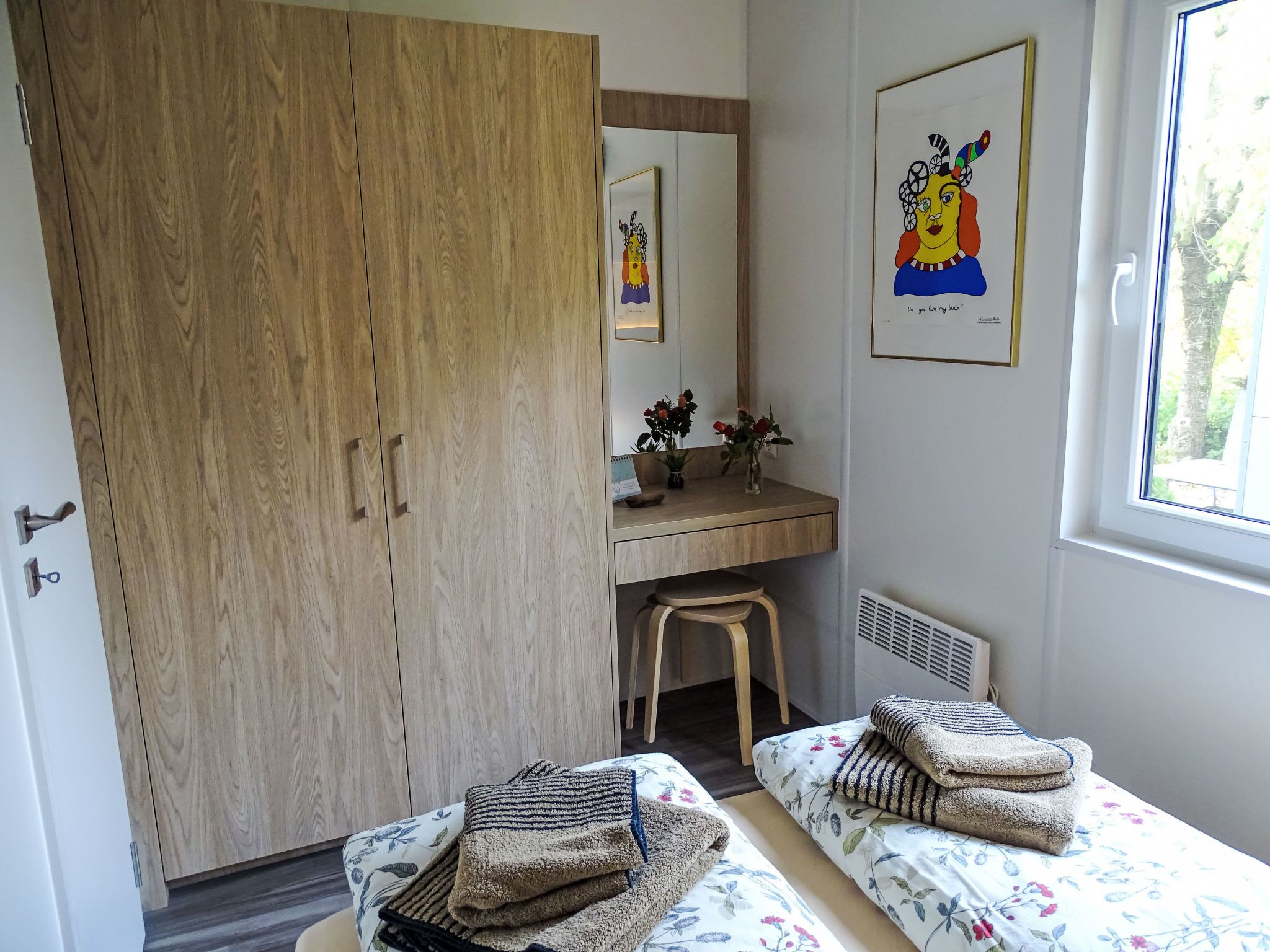 Une chambre avec une armoire en bois, un bureau avec un miroir, deux lits avec une literie à motifs floraux et une fenêtre donnant sur des arbres.