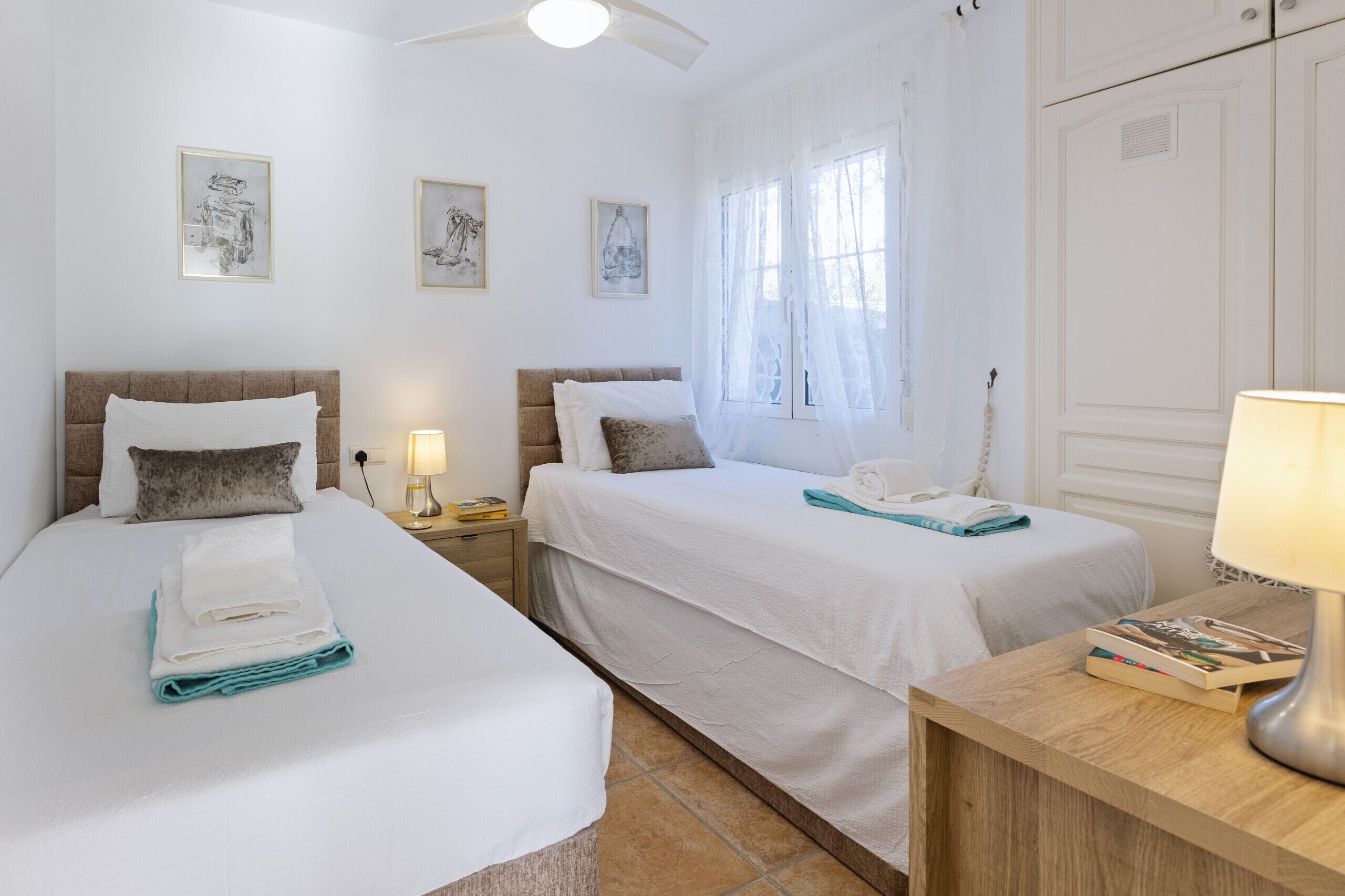 A hotel room with two beds, a wooden nightstand with a lamp, and a window with sheer curtains.