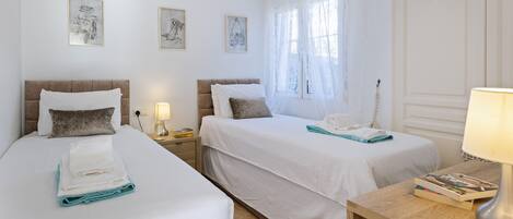 A hotel room with two beds, a wooden nightstand with a lamp, and a window with sheer curtains.