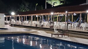 Un área de piscina nocturna con sillones y mesas blancas bajo una pérgola.