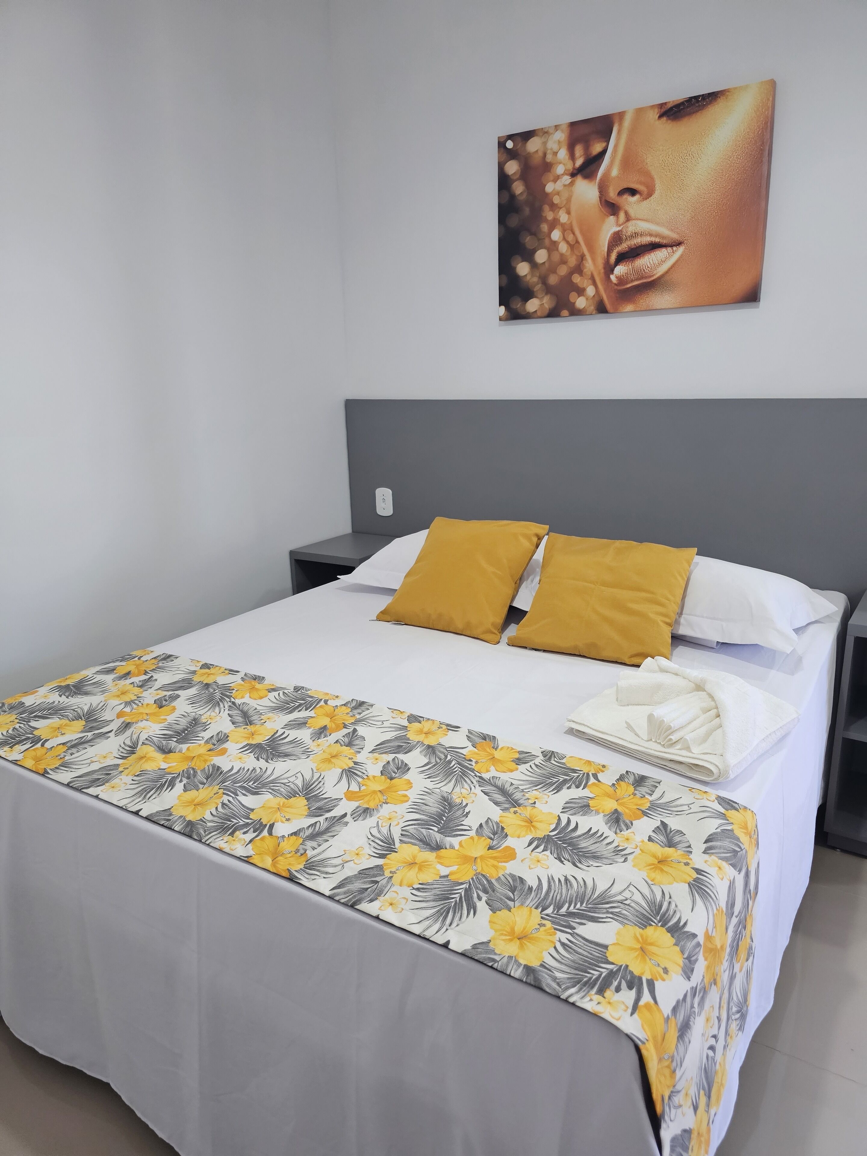 Una cama impecable con un edredón floral, dos almohadas color mostaza y una toalla blanca doblada al lado.