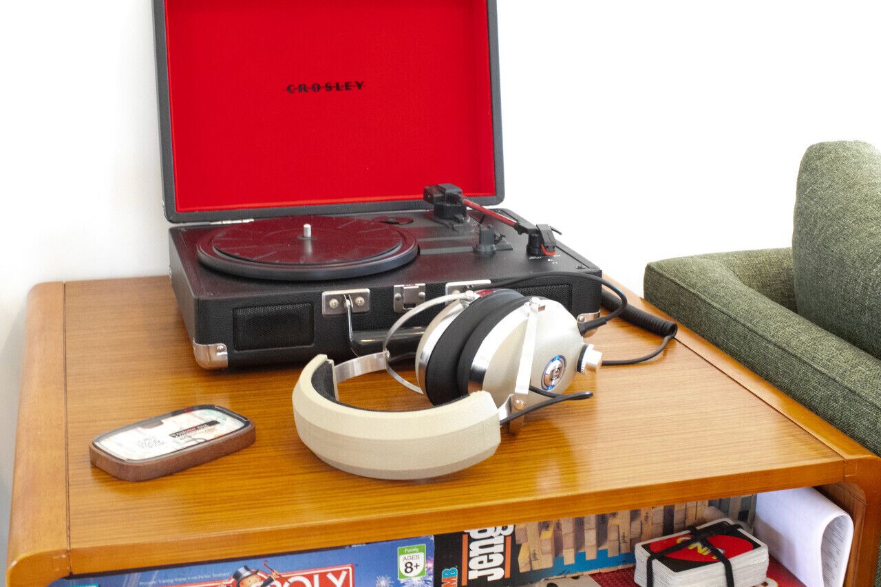 Tocadiscos con auriculares sobre una mesa de madera, un estuche rojo y una silla verde.
