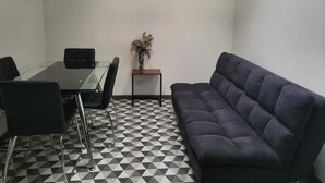 Una habitación con una mesa de vidrio, sillas negras, un sofá oscuro, una mesita auxiliar con un florero y un suelo a cuadros blanco y negro.