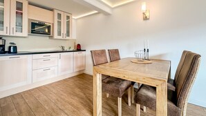 A modern kitchen with white cabinets, a built-in oven, and a dining area with a wooden table and leather chairs. - Appartement Disüdlöner (Sylt)