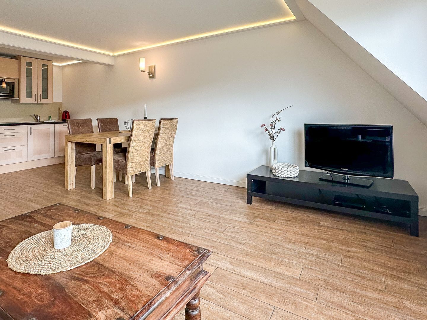 A modern living room with a wooden dining table, chairs, a TV on a black stand, and a kitchen in the background.