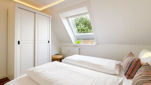 A bedroom with a skylight, a bed with white bedding, a wooden stool, and a radiator. - Appartement Disüdlöner (Sylt)