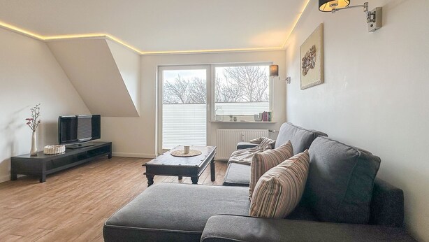 A modern loft apartment with a sloped ceiling, a grey sofa, a small coffee table, a TV on a stand, and a large window with blinds. - Appartement Disüdlöner (Sylt)