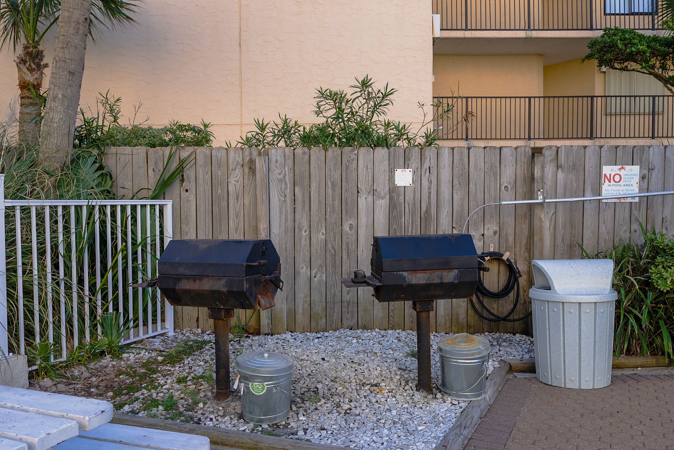 Twee buitenbarbecuegrills, een houten omheining en een vuilnisbak.
