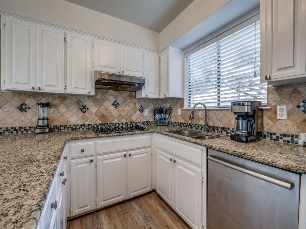A kitchen with white cabinets, a granite countertop, a stainless steel dishwasher, and a window with blinds.