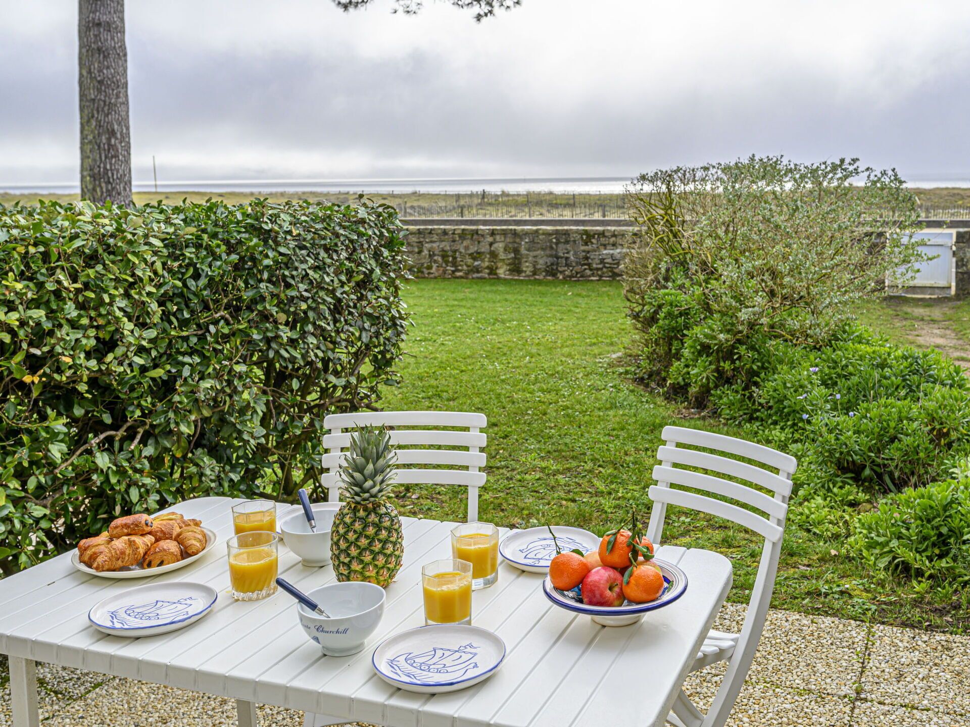 Una mesa exterior con alimentos para el desayuno, como cruasanes, piña, naranjas y jugo, con un jardín y un muro de piedra de fondo.