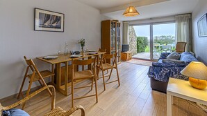 Zona pranzo con tavolo in legno apparecchiato per quattro persone, televisione su mobiletto in legno e ampia finestra con vista su vegetazione e un edificio.