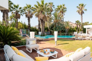 Un espace aménagé au bord de la piscine, avec des chaises longues, une table à manger où sont servis des plats et des boissons, et une piscine entourée de palmiers.