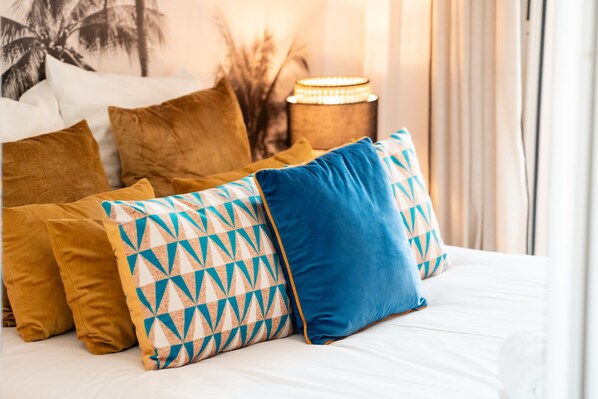 A bed with multiple pillows, a nightstand with a lamp, and a wall with a tropical-themed painting.