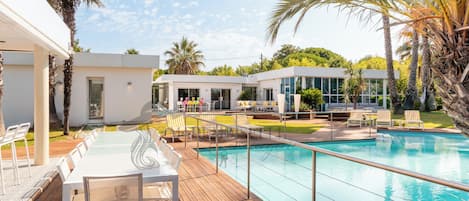 A pool area with a wooden deck, lounge chairs, and a modern building with large windows.