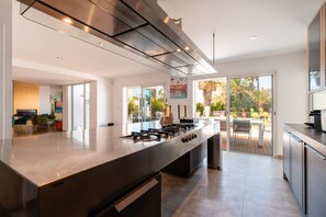 Une cuisine moderne dotée d’un grand îlot central, d’appareils électroménagers en acier inoxydable et offrant une vue sur la terrasse extérieure.
