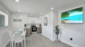 A modern kitchen with white cabinets, a dining area with white chairs, a TV mounted on the wall, and a beach scene on the wall. - Walk to Beach 2BR | Bright Pompano Retreat (Pompano Beach)
