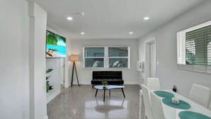 A modern living room with a black sofa, a coffee table, and a dining area with white chairs and a table. - Walk to Beach 2BR | Bright Pompano Retreat (Pompano Beach)