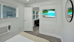 A bedroom with a bed, a window with blinds, a door, a wall-mounted TV, and a mirror. - Walk to Beach 2BR | Bright Pompano Retreat (Pompano Beach)