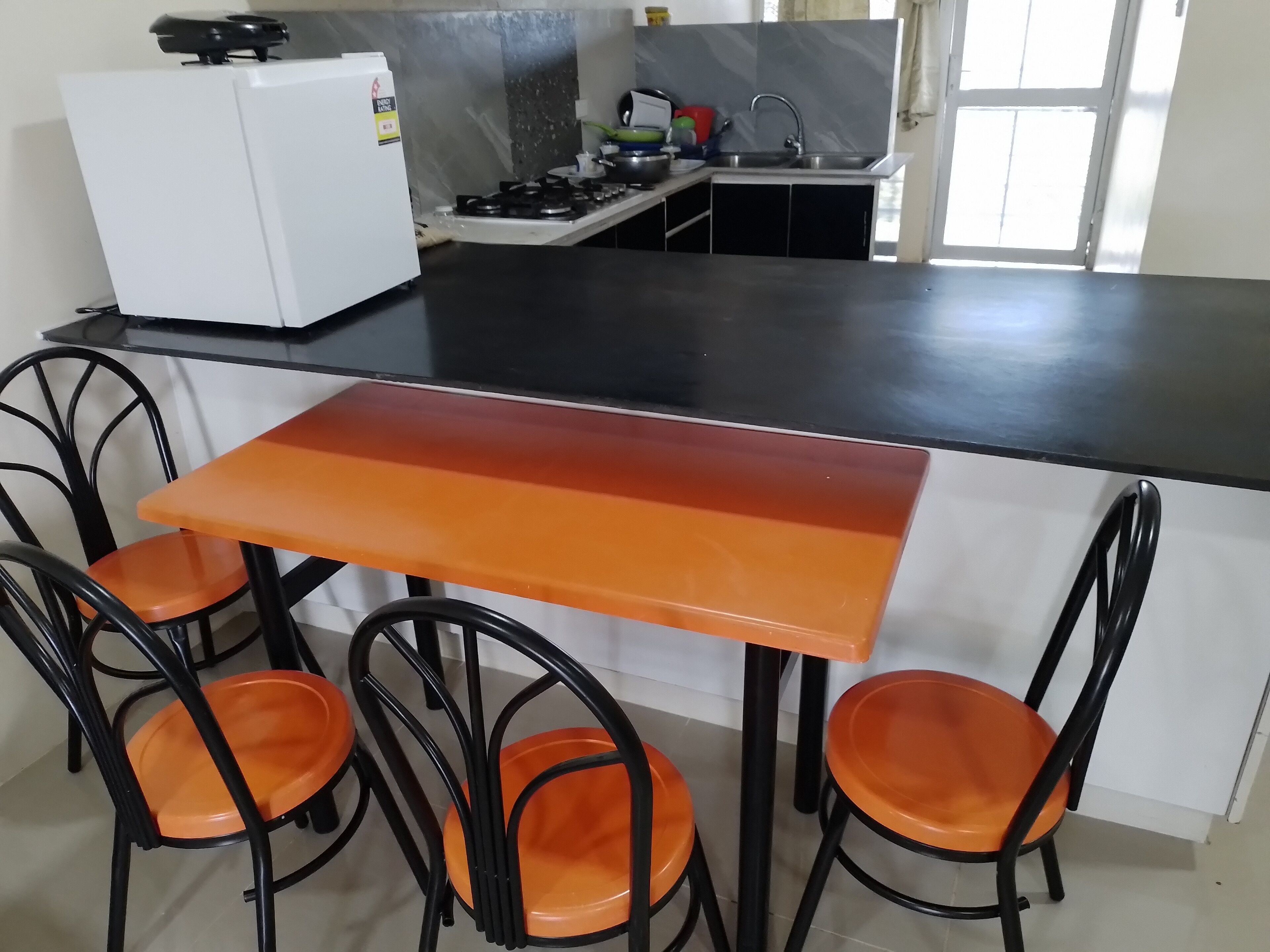 Et kompakt køkken med en sort bordplade, hvide skabe og en lille vask. Fire orange stole står ved et bord.