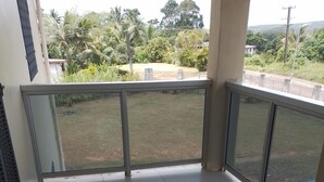 Un balcón con barandilla de cristal, suelo de baldosas y vista a un área verde con árboles y un edificio al fondo.