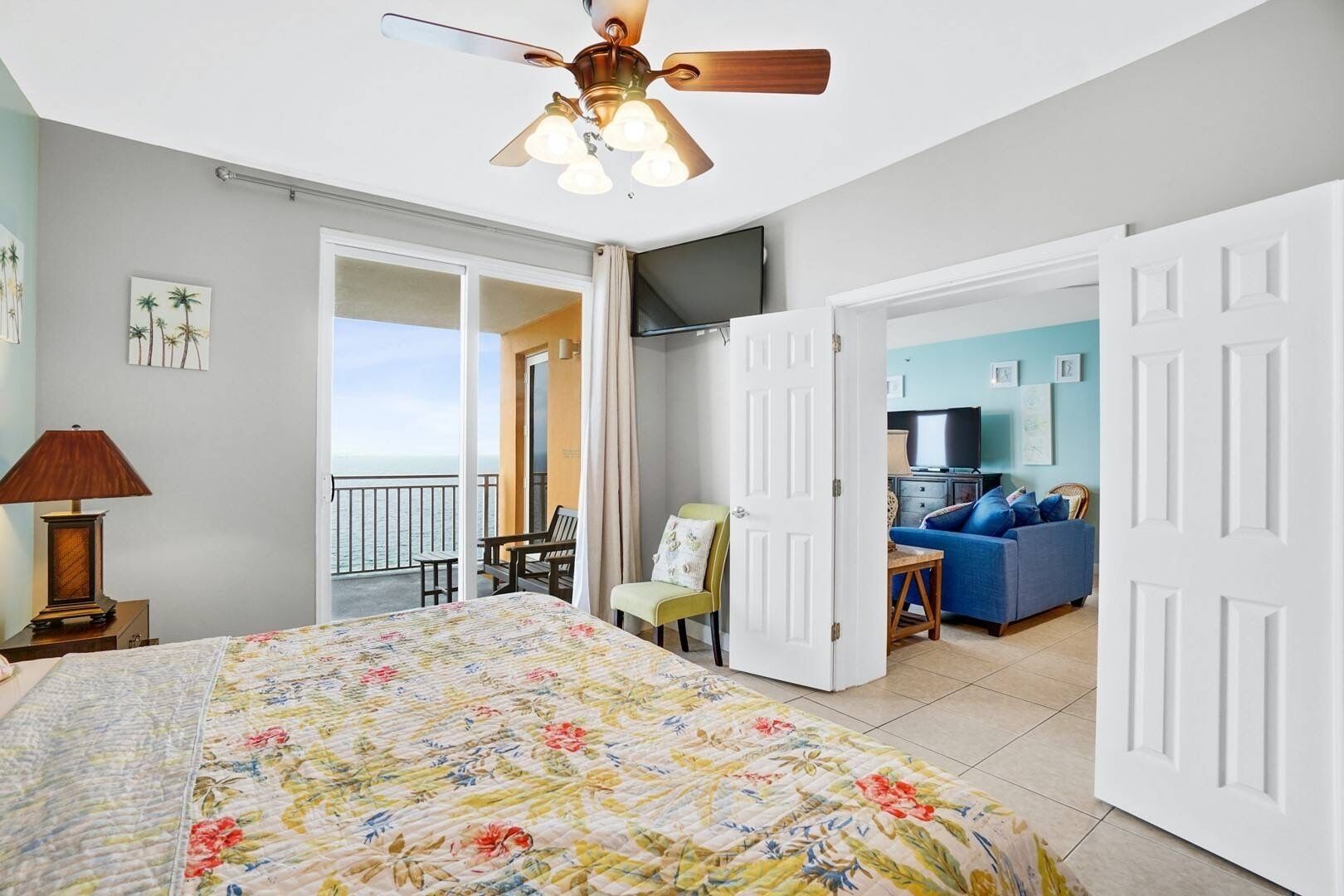 A bedroom with a bed, a ceiling fan, a nightstand, a lamp, a balcony with a view of the sea, and a living area with a sofa and a TV.
