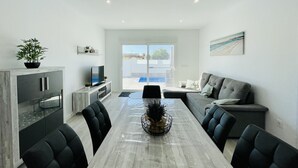A modern living room with a grey sofa, a dining table, and a TV on a cabinet. There is a balcony with a pool view. - Villa Tranquilidad (Avileses) (Avileses)