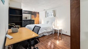 Ein Schlafzimmer mit Bett, Schreibtisch mit Stuhl, einem kleinen Tisch mit Lampe, einem Kleiderschrank und einer Wand mit Palmenmotiv.