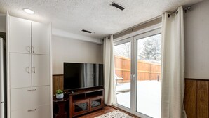 Ein Wohnzimmer mit Flachbildfernseher, Holzboden, einem weißen Schrank und einer Glastür zum Garten.