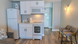 A compact kitchen with white cabinetry, a microwave, a stove, and a refrigerator. There are two bar stools and a sofa in the adjacent living area. - Sea Mist 20407 (Myrtle Beach)