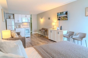 A compact hotel room with a bed, a desk, a kitchenette, and a seating area with chairs.
