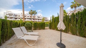 A hotel courtyard with two lounge chairs and a single umbrella, surrounded by neatly trimmed hedges and palm trees.