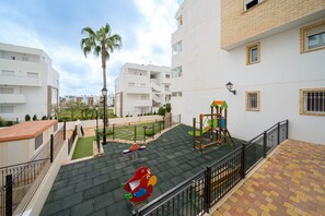 A playground with a slide and swings, surrounded by a fenced area with a palm tree and a tennis court. - Apartamento Mariphils (Linnea Sol) (Orihuela Costa)