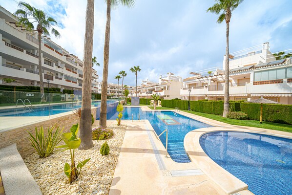 A swimming pool with a curved design, surrounded by palm trees and a well-maintained hedge. - Apartamento Mariphils (Linnea Sol) (Orihuela Costa)