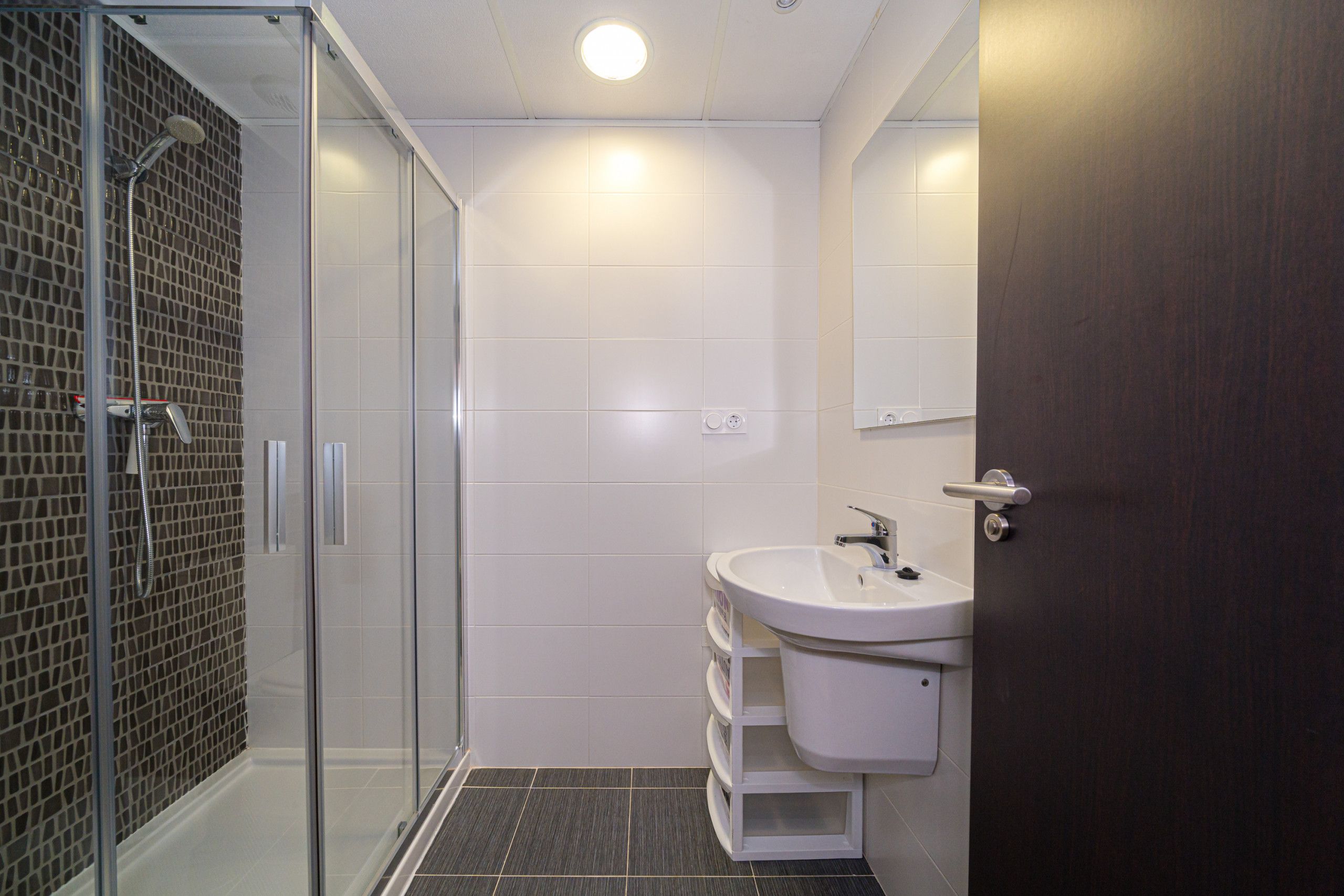A modern bathroom with a glass shower enclosure, a white sink, and a dark tiled shower wall.