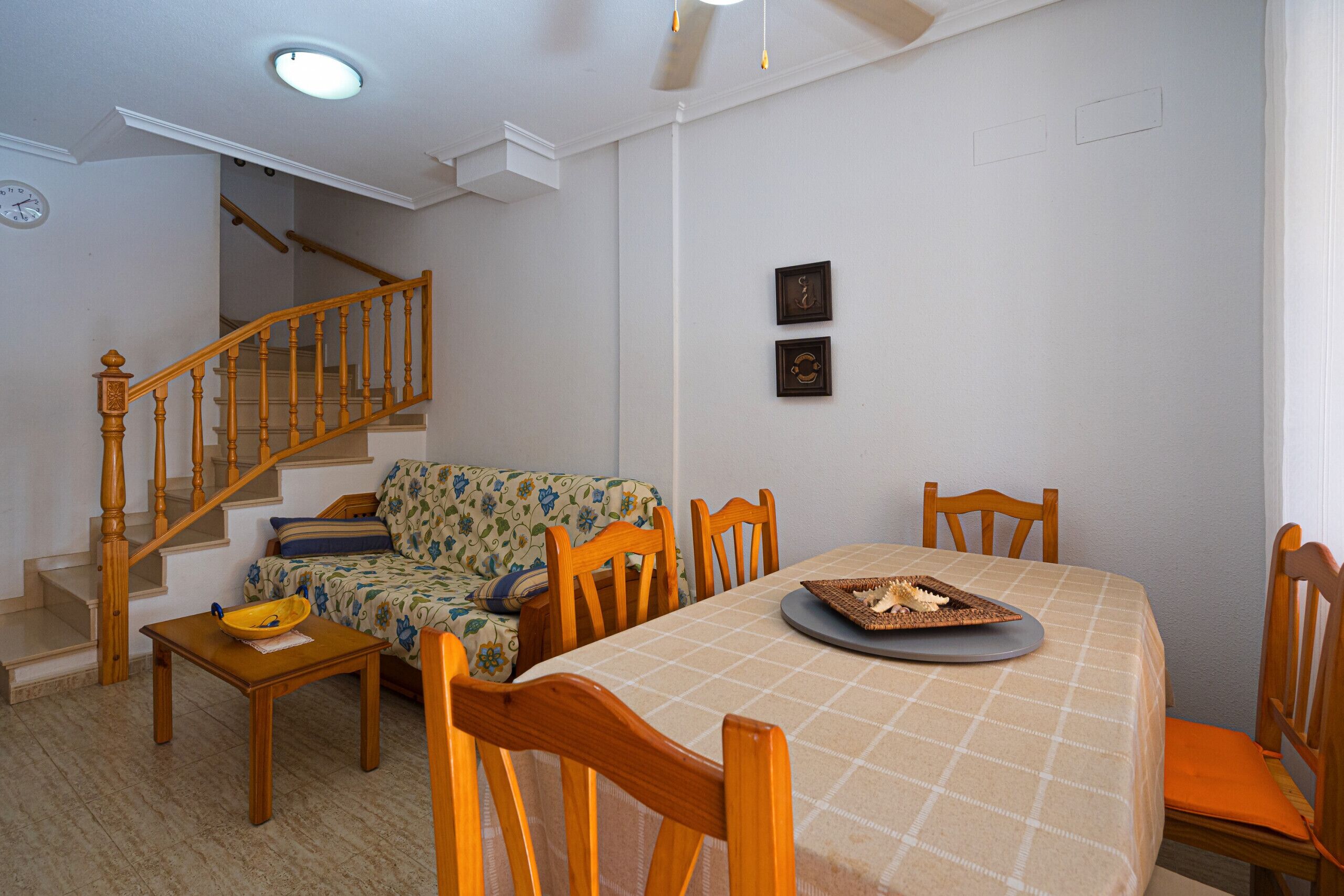 Ein Essbereich mit einem Tisch für vier Personen, Holzsesseln, einer Treppe und einer mit Blumenmuster bezogenen Couch.