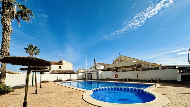 Une piscine avec une terrasse carrelée, des palmiers et, au fond, un bâtiment dont le toit est également recouvert de tuiles.