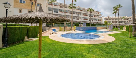A swimming pool with a thatched roof structure, surrounded by well-maintained grass and palm trees, with a multi-story building in the background.
