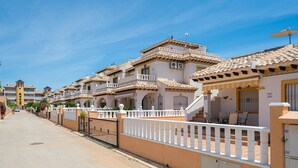 Une rangée de bâtiments traditionnels de style méditerranéen, dotés de balcons et de toits en tuiles, une entrée fermée par un portail et un ciel dégagé.